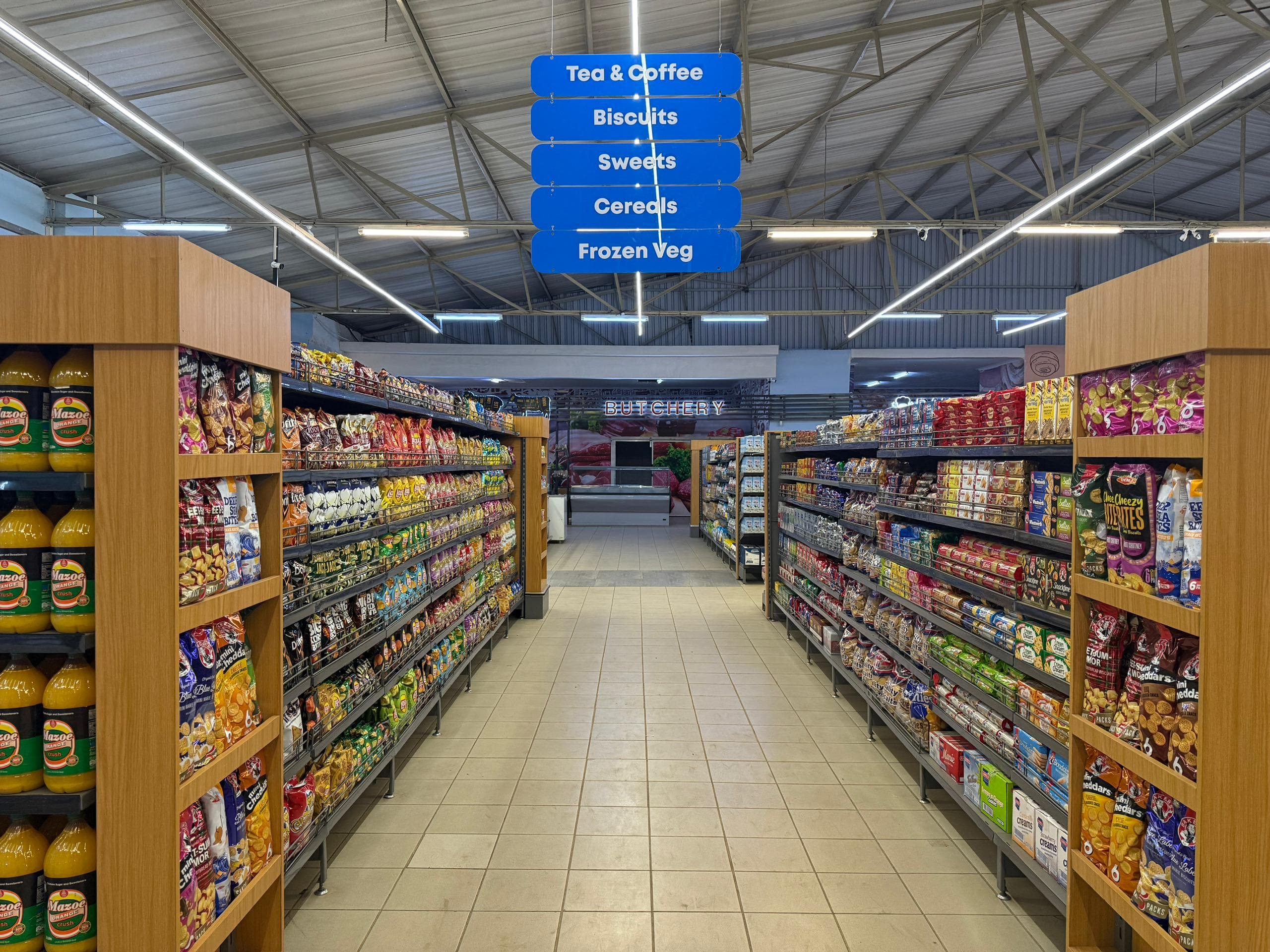 An aisle inside the PrevailMart Mazowe branch, stocked with groceries under overhead category signs.